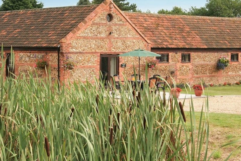 Rustic brick cottage with a tile roof seen through tall garden reeds; patio seating under a green umbrella in front of the entrance.