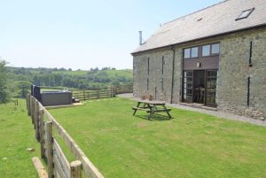 Stone cottage with glass doors opening to a green lawn, a wooden picnic table, and distant rolling countryside under a clear blue sky.