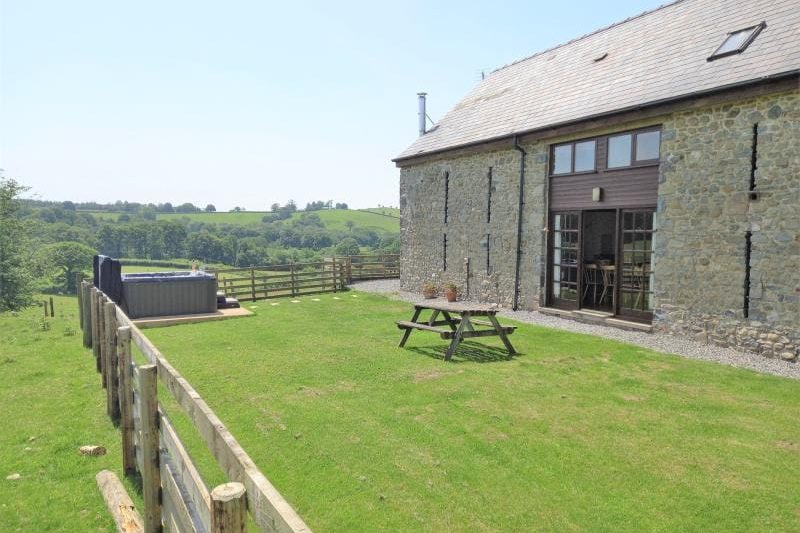Stone cottage with glass doors opening to a green lawn, a wooden picnic table, and distant rolling countryside under a clear blue sky.