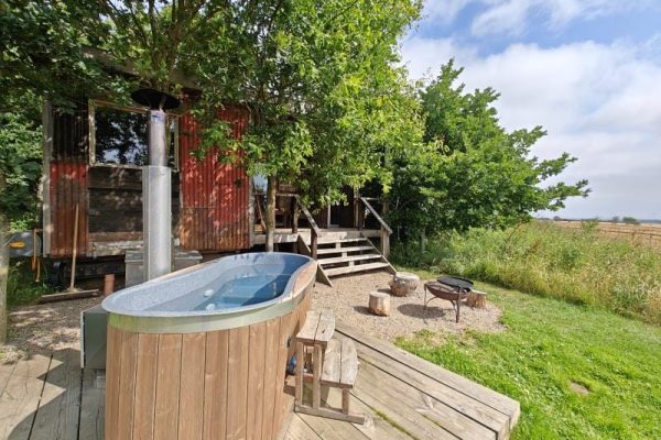 Wooden deck with an outdoor hot tub beside a rustic red cabin, surrounded by trees and a grassy yard under a blue sky.