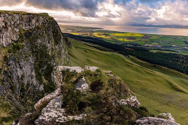 Steep rocky cliff with scrubby vegetation overlooking green fields and a distant coastline.