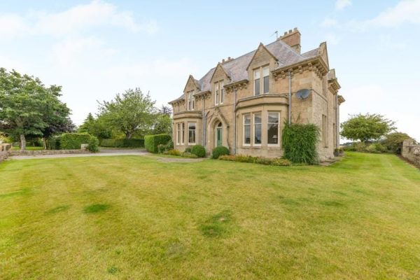 Stone Victorian-style house with arched doorway, surrounded by a manicured lawn and trees in a rural setting