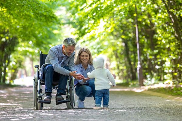 Man in a wheelchair and a woman kneel on a sunlit park path, smiling as a toddler in a white hoodie approaches to greet them.