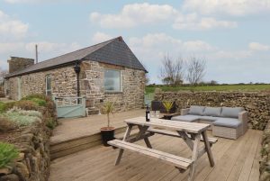 Stone cottage with a slate roof on a wooden deck, outdoor dining table and seating area against a stone wall backdrop.