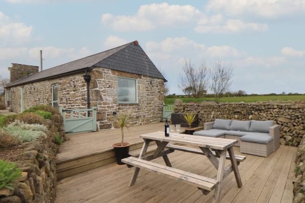 Stone cottage with a slate roof on a wooden deck, outdoor dining table and seating area against a stone wall backdrop.