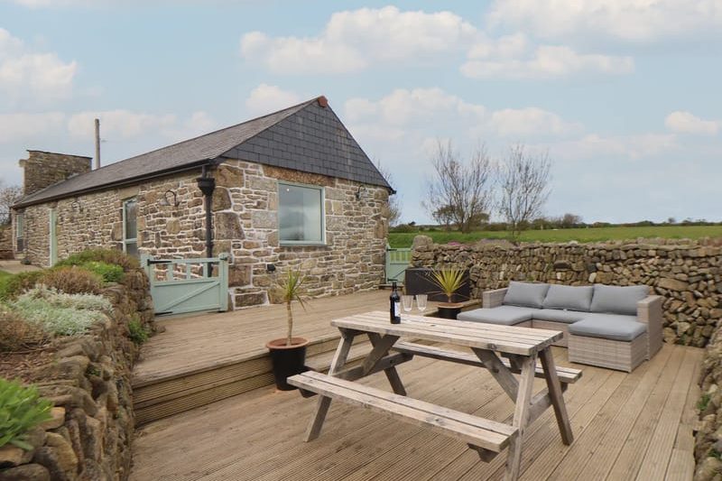 Stone cottage with a slate roof on a wooden deck, outdoor dining table and seating area against a stone wall backdrop.