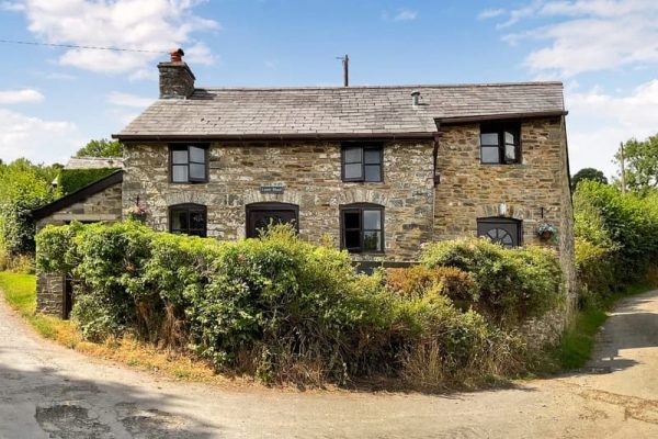 Two-story stone cottage at a rural crossroads, with a slate roof and dark-framed windows, surrounded by tall hedges and a dirt road.