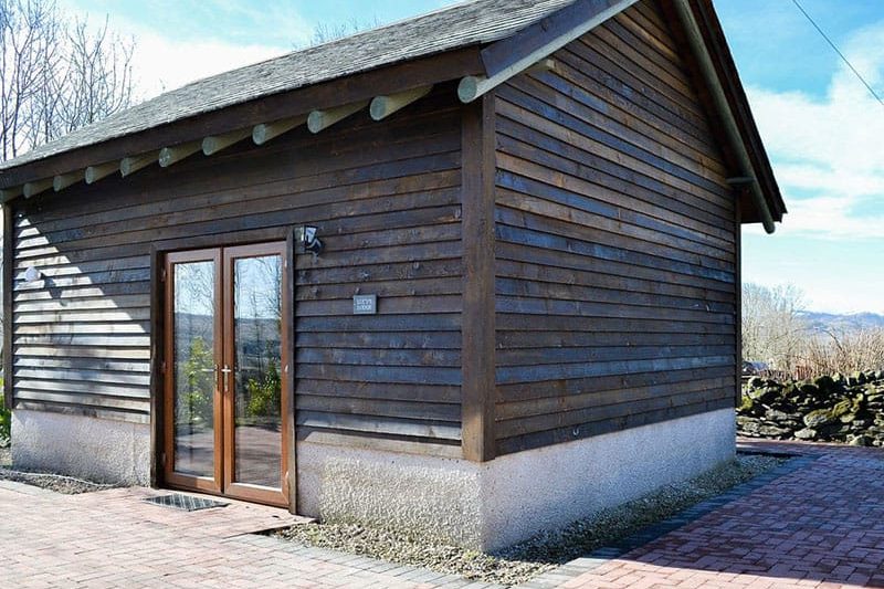 Wooden cabin with dark stain, glass double doors, and a concrete base on a brick-paved path, set in a rural area.
