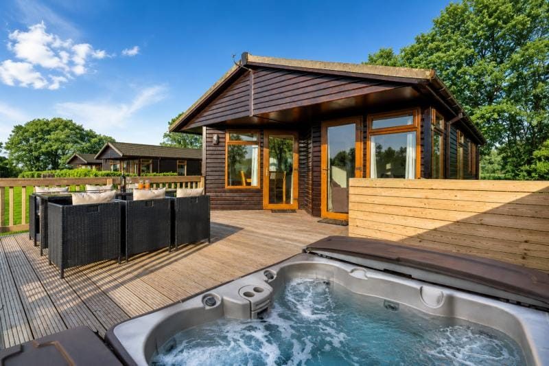 Wooden cabin on a spacious deck with a bubbling hot tub, outdoor seating, and glass doors, surrounded by trees under a blue sky.