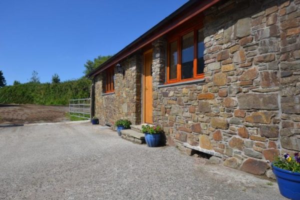 Stone cottage with orange-framed windows and a wooden door, blue flower pots along a gravel driveway under a blue sky.