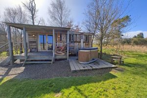 Rustic wooden cabin with a small deck, lounge chairs, and a circular outdoor hot tub on a raised platform in a rural yard.