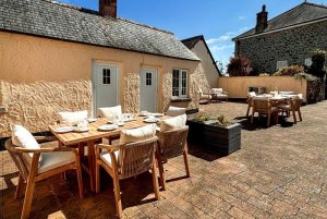 Outdoor patio with a wooden dining table and cream cushions beside a peach stucco cottage on a sunny day nearby stone buildings.