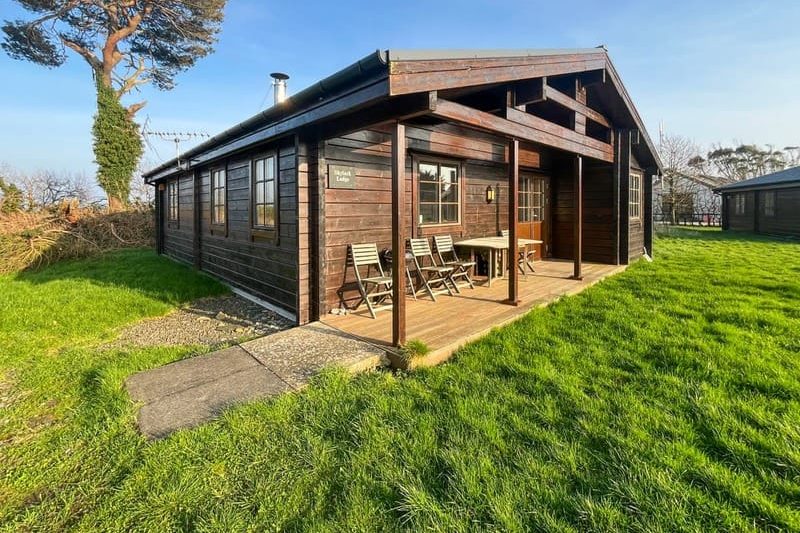Wooden cabin with a covered porch and outdoor seating on a deck, set in a grassy yard under a blue sky
