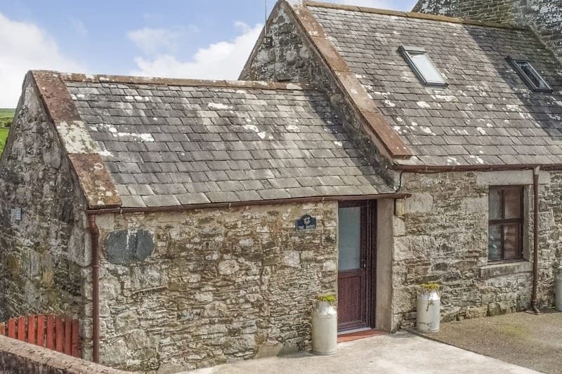 Stone cottage with a slate roof, wooden door, and a window; two gas cylinders stand by the entrance.