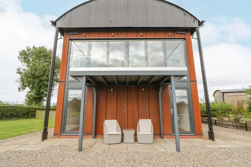 Modern two-story glass-and-wood house with a shaded patio and two beige chairs around a small table on a stone-paver patio