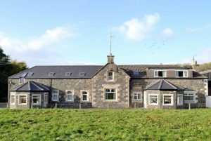Long stone farmhouse with a slate roof, chimneys and bay windows, set in a green grassy field under a blue sky.