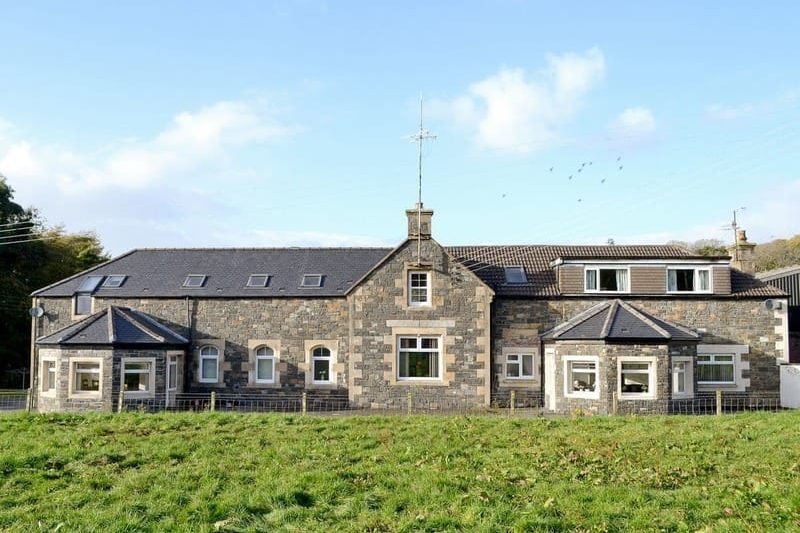 Long stone farmhouse with a slate roof, chimneys and bay windows, set in a green grassy field under a blue sky.