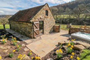 Stone cottage with a red-tiled roof, open wooden door, and stone-paved patio amid yellow daffodils in a sunny garden. (informative)