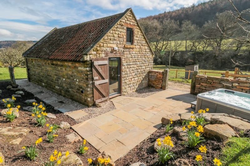 Stone cottage with a red-tiled roof, open wooden door, and stone-paved patio amid yellow daffodils in a sunny garden. (informative)