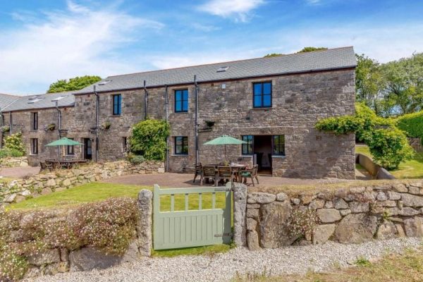 Stone two-story stone cottage with blue-framed windows, outdoor dining area under green umbrellas on a gravel patio behind a low stone wall.