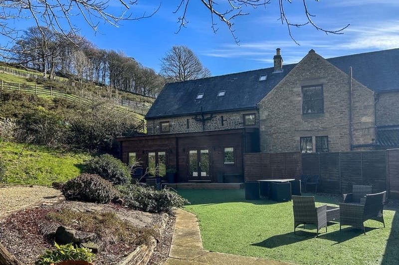 Stone house with slate roof and a wooden extension, backdrop of a hillside and blue sky, with a manicured lawn and outdoor seating area in the courtyard.