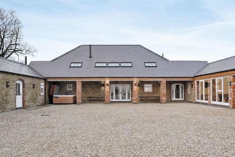 Wide view of a gravel courtyard in front of a brick-and-stone house with a central covered patio and skylit roof panels behind columns.