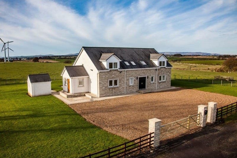 White stone house with gray roof and gravel driveway on a rural farm, wind turbine in the background.